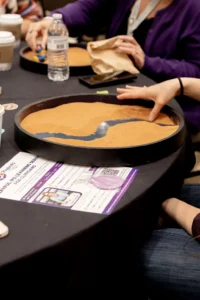 A hand carving a river channel through orange sand in a round tray