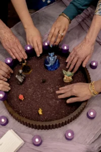 Many hands reaching toward one round tray of dark sand ringed with tealights and small figures