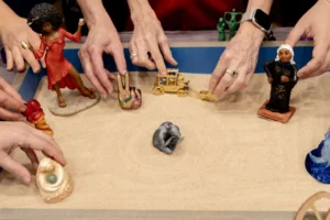 Several hands placing miniatures, among them a figure in a red dress and a gold carriage, into a pale sandtray