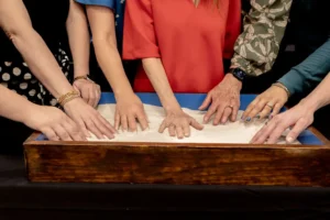 Seven pairs of hands smoothing white sand side by side in one long wooden tray