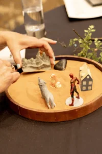 Fingers lowering a mountain piece into a round tray beside a wolf and a small house
