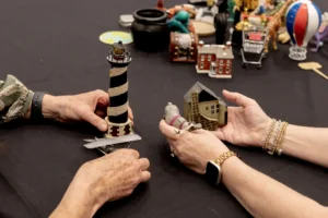 Two pairs of hands passing a striped lighthouse and a cottage miniature across a table
