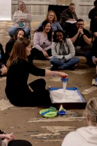 A presenter seated on the floor pouring sand into a tray while a circle of therapists watches
