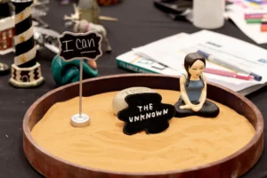 A round tray of warm sand holding a small flag reading I can, a black stone and a seated figure