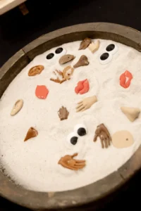 A round tray of white sand scattered with small sculpted eyes, lips, ears and hands