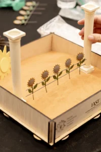 A wooden IAST sandtray where two white columns frame a row of sunflowers as a hand sets one column